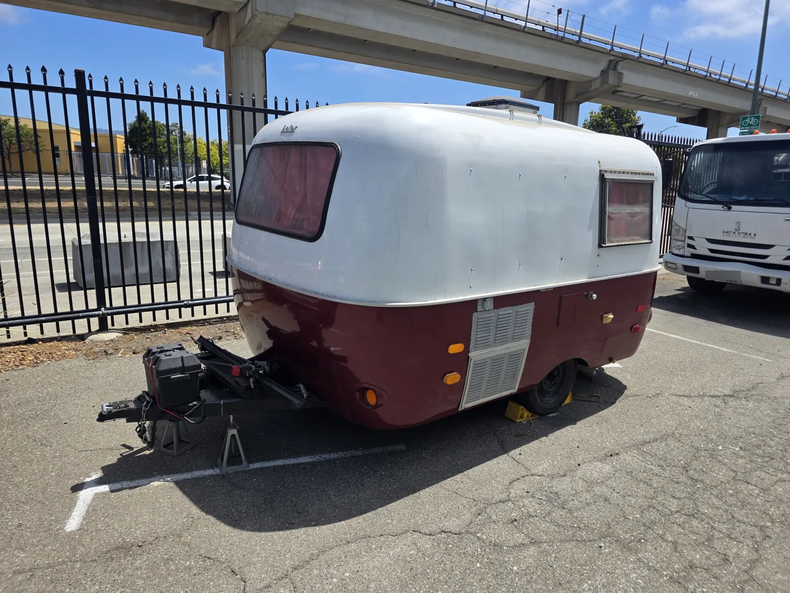 Side view of a vintage Boler fiberglass camper trailer on jack stands in a parking lot beneath a freeway.