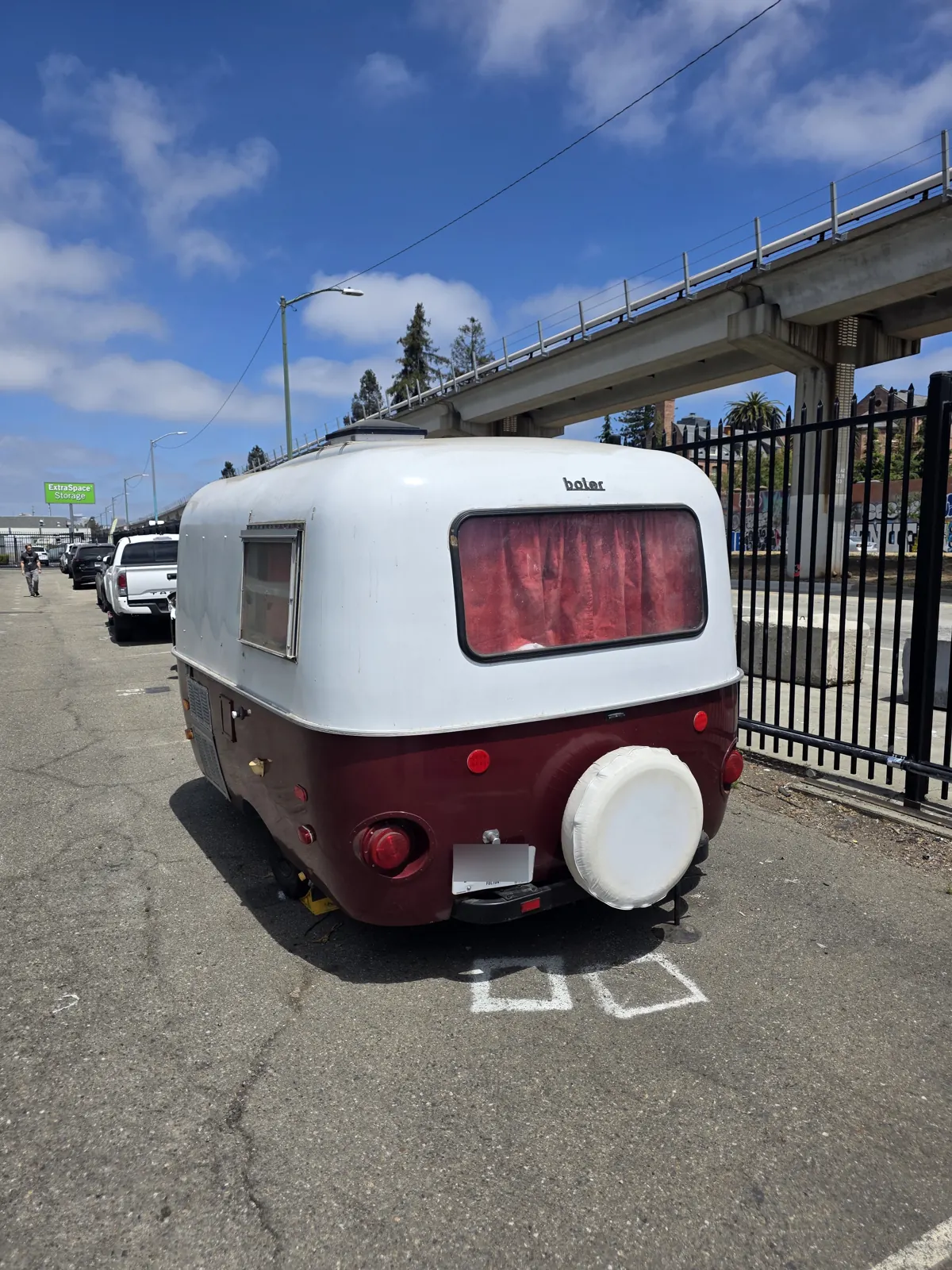 Rear view of a vintage Boler camper trailer with a spare tire cover, parked under an elevated rail line.