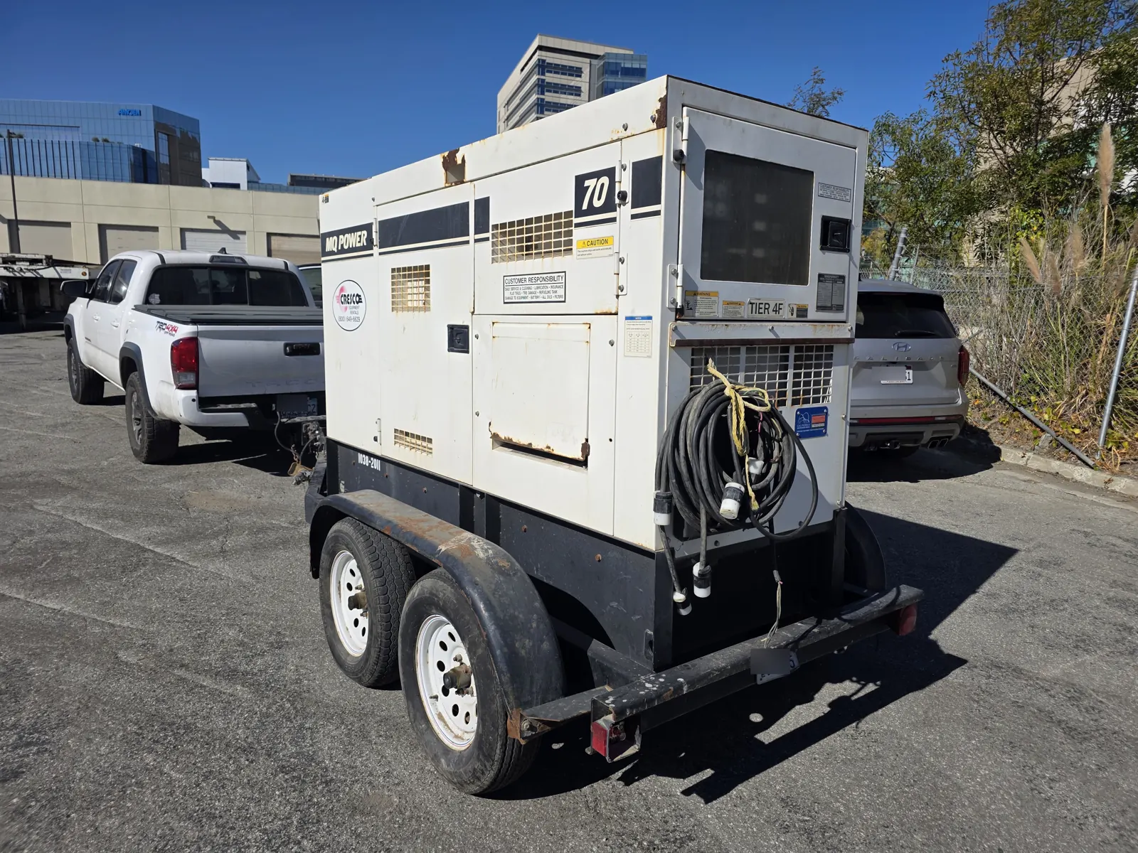 Rear three-quarter view of a towable generator trailer with power cables coiled on the back, a pickup truck in front.