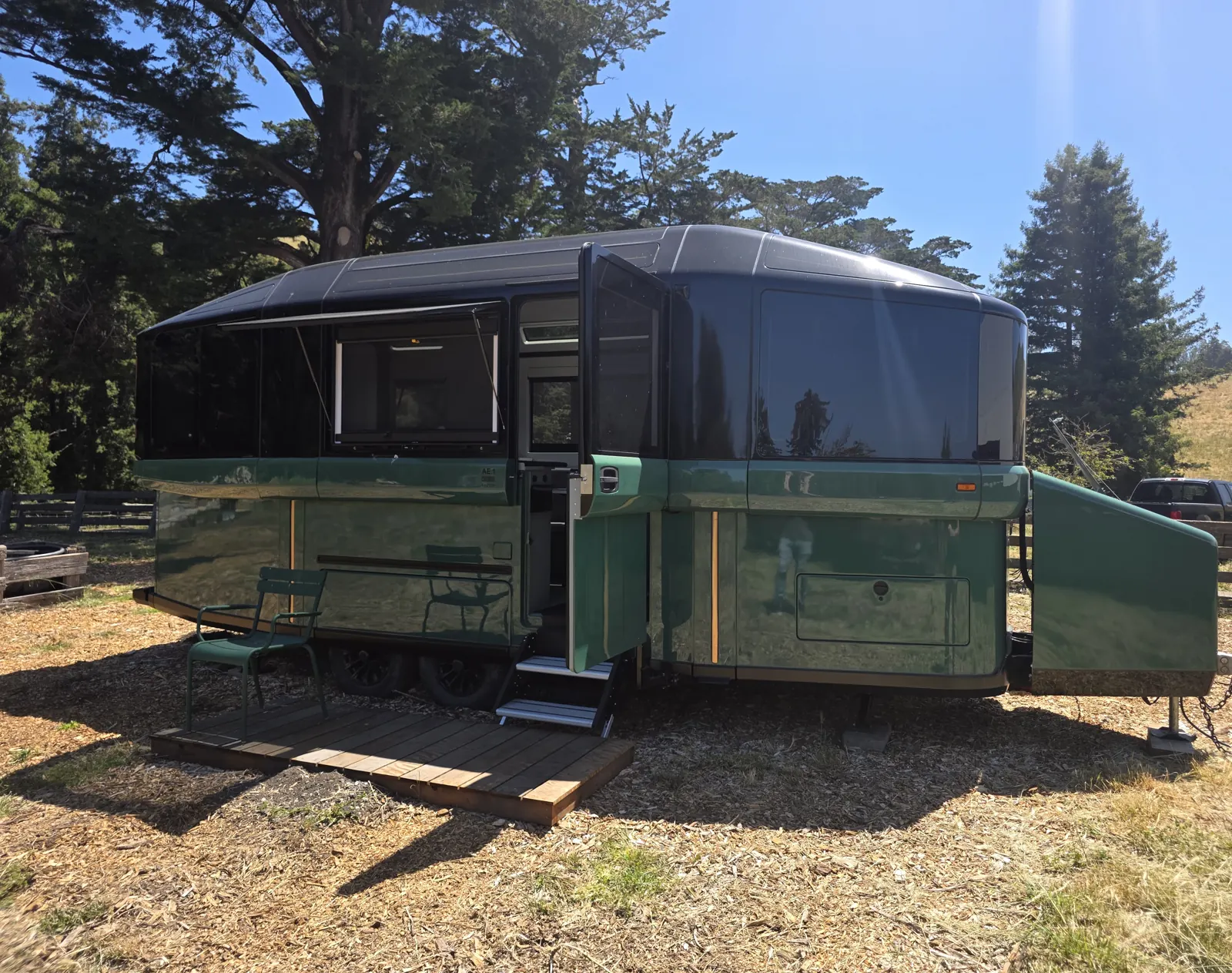 Green Lightship L1 electric travel trailer with its roof raised, parked on a ranch with a wooden deck and chairs out front.