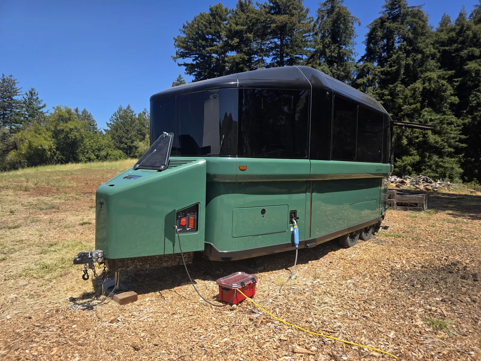 Front three-quarter view of a green Lightship L1 trailer with its charge cable plugged in, redwoods behind.