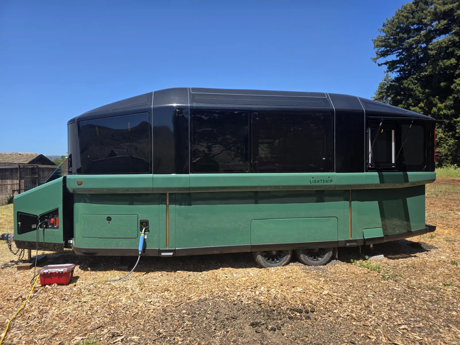 Side profile of a green Lightship L1 electric trailer with dark glass upper body, parked on wood chips beside a barn.