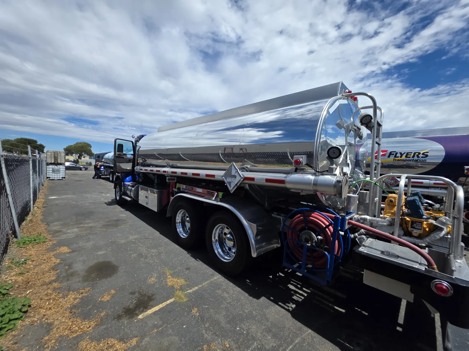 Polished stainless steel fuel tanker truck with hose reels, parked in a fenced yard under a cloudy sky.
