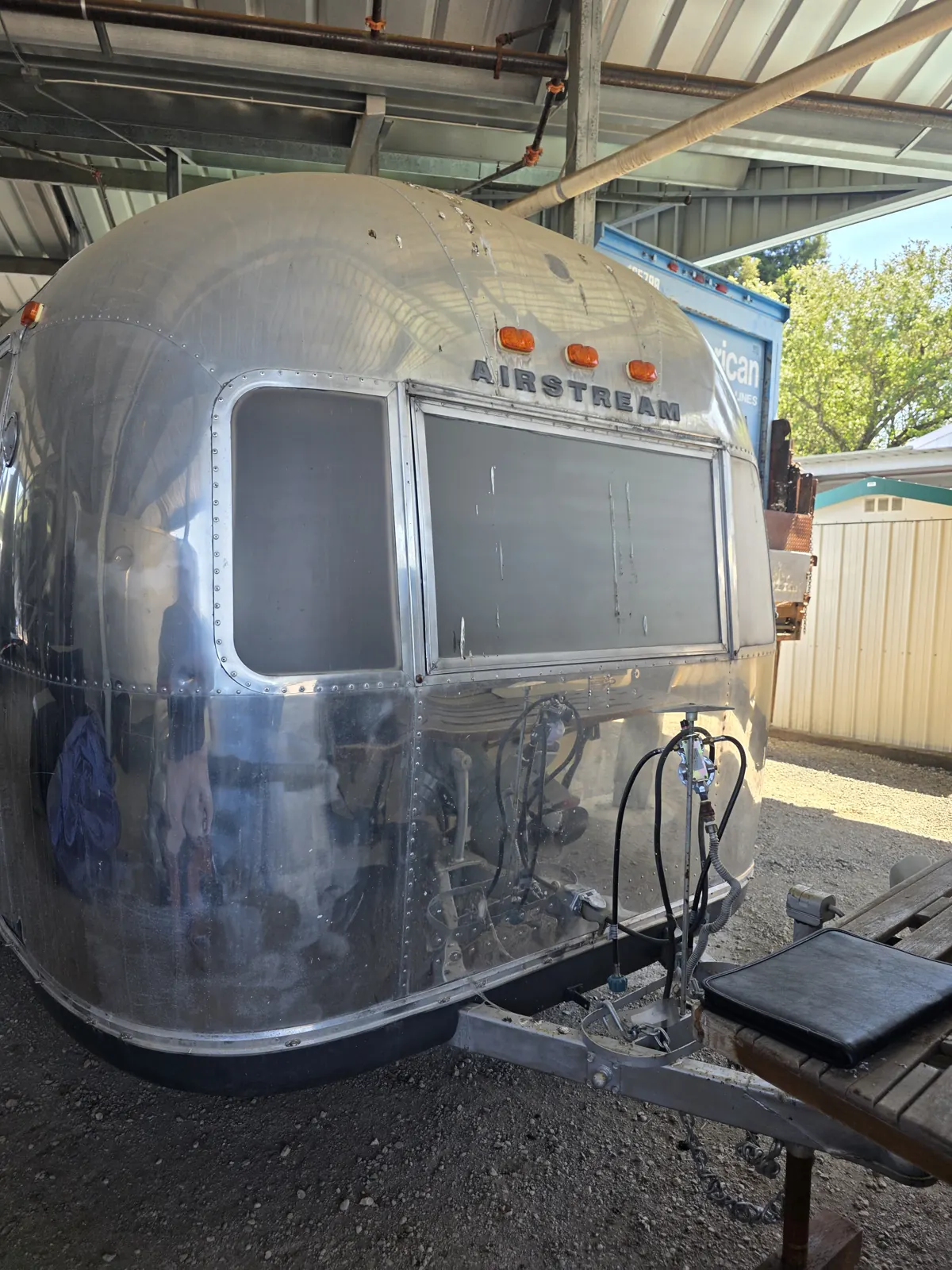 Front view of a vintage polished-aluminum Airstream trailer with propane tanks on the tongue, parked under a metal canopy.